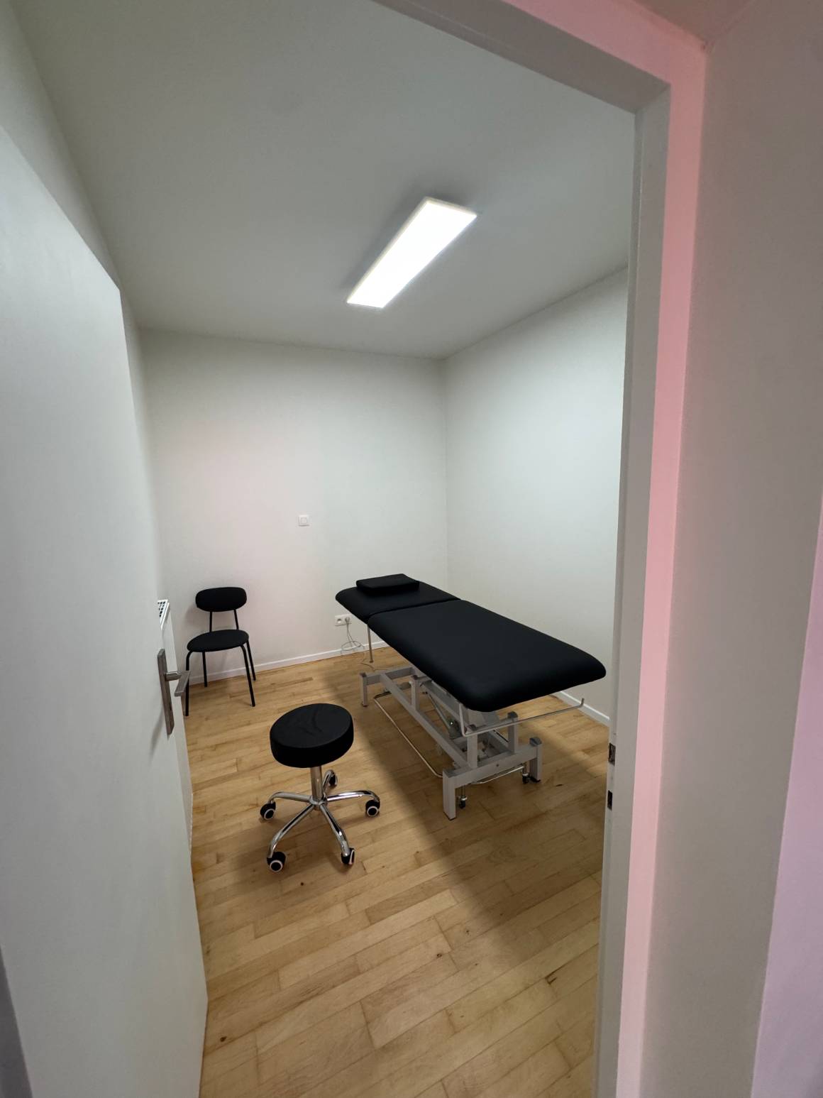 Un cabinet privé du centre KINE+ vu depuis le couloir : table de soin, tabouret et chaise sur parquet clair