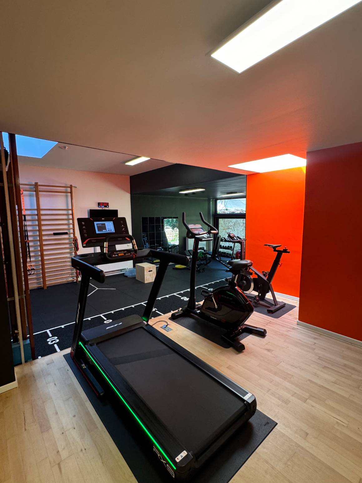 La zone cardio du centre KINE+ : tapis de course et vélo devant le mur orange, espaliers et miroir sur la gauche