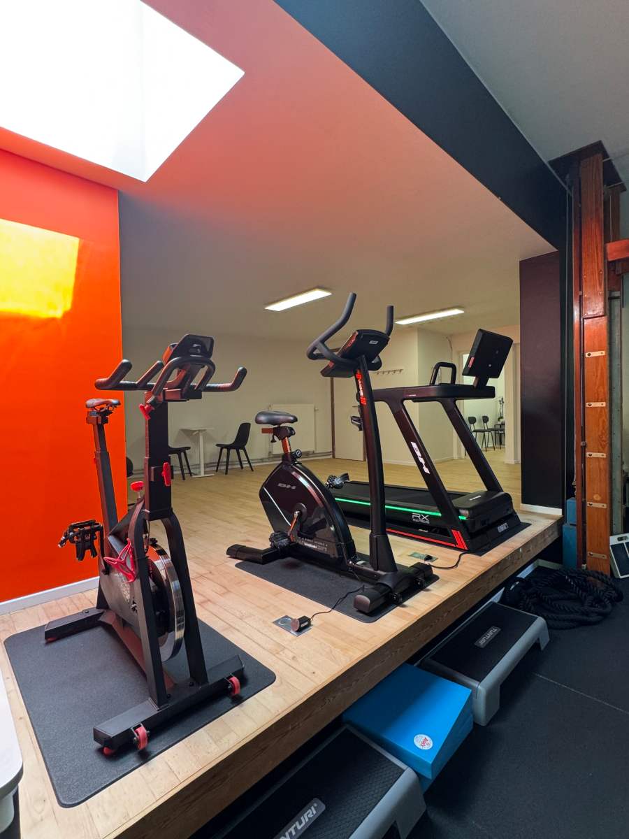 La zone cardio du centre KINE+ : vélos et tapis de course sur l'estrade, devant le mur orange